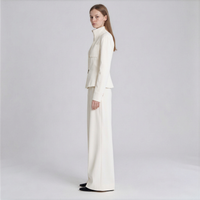 A woman stands in profile wearing a matching cream-colored tailored set consisting of a structured long-sleeve jacket with a high mock neck and wide-leg trousers, photographed against a light gray studio background.