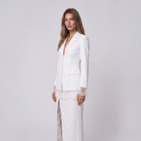 A woman modeling a white tailored blazer with lace trim at the cuffs and hem, paired with a matching white skirt, against a plain light gray studio background.